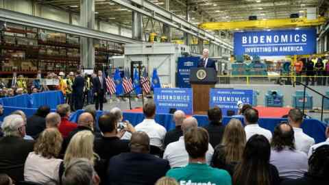 A man in suit stands on a podium inside a factory and addresses a crowd of people