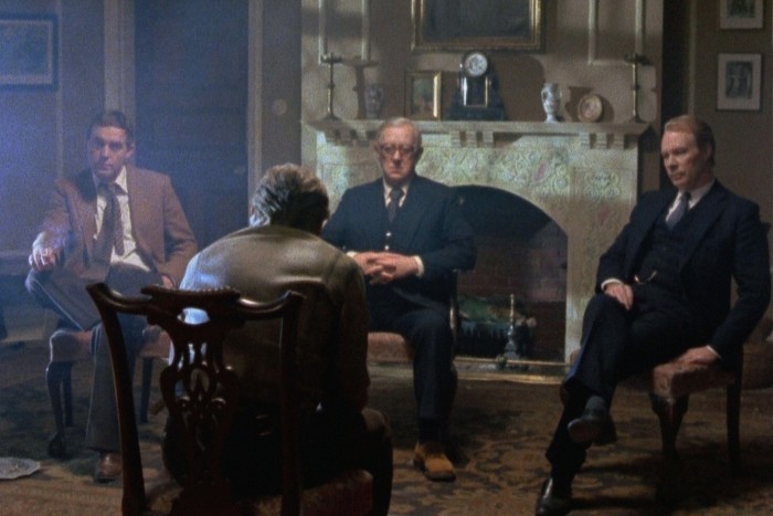 Four men in suits sit on chairs, facing each other, in a rather old-fashioned drawing room  