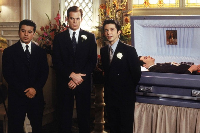 Three men in dark suits stand in front of any open casket in a funeral parlour